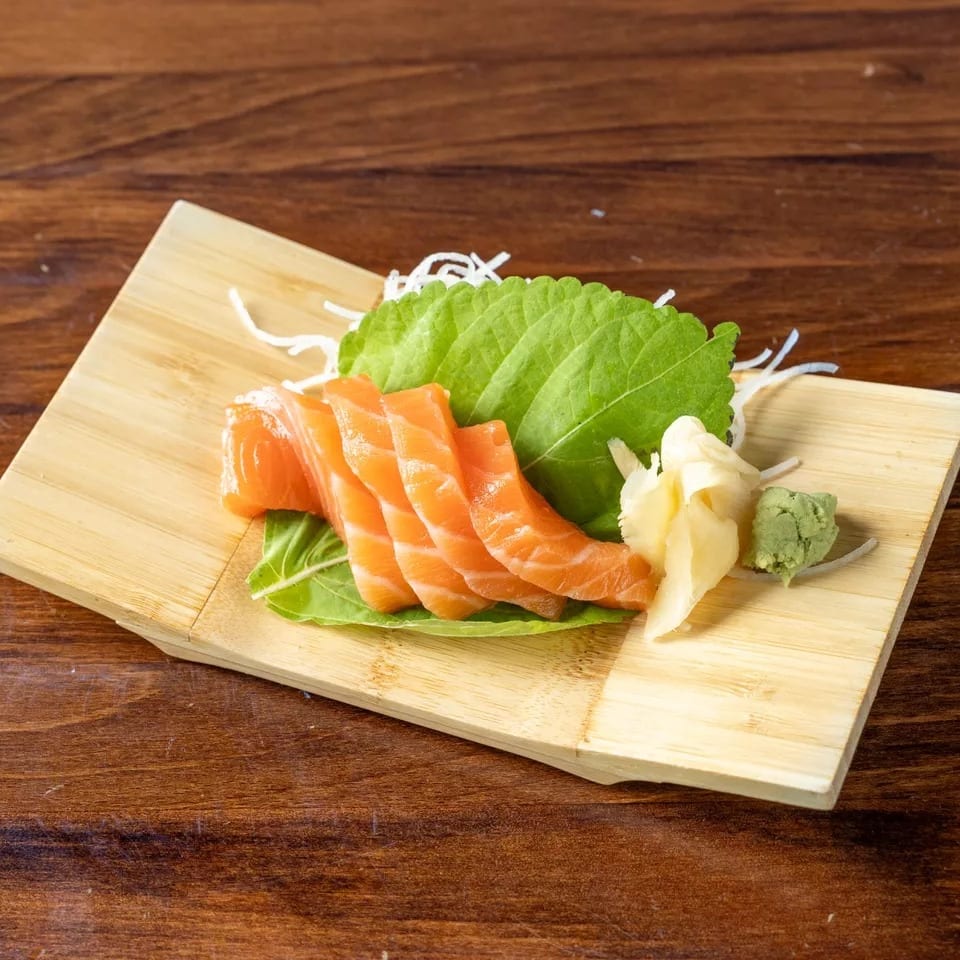 Fresh salmon sashimi slices with ginger, wasabi, and shiso leaf on a wooden board.