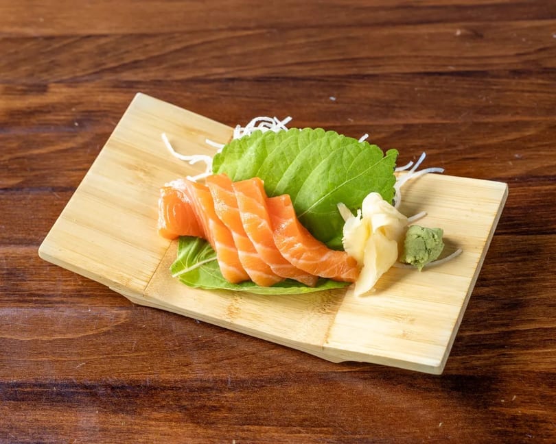 Fresh salmon sashimi slices with ginger, wasabi, and shiso leaf on a wooden board.