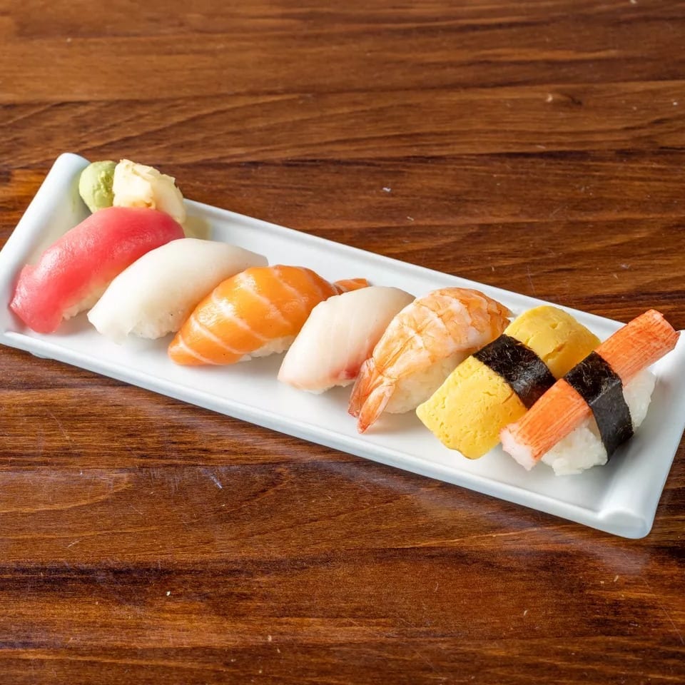 A white rectangular plate holds a variety of colorful nigiri sushi, with wasabi and ginger.
