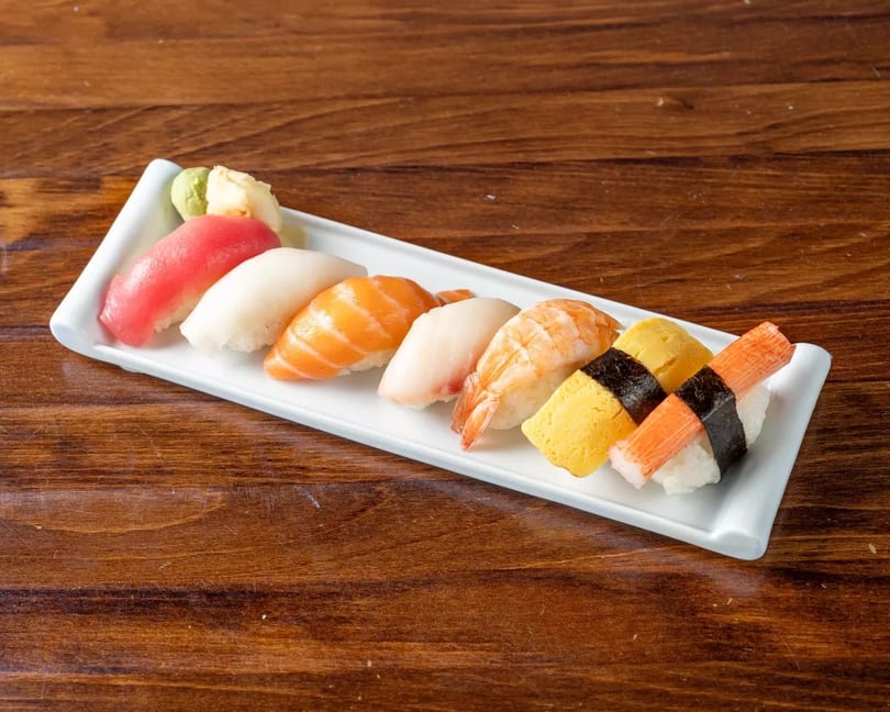 A white rectangular plate holds a variety of colorful nigiri sushi, with wasabi and ginger.