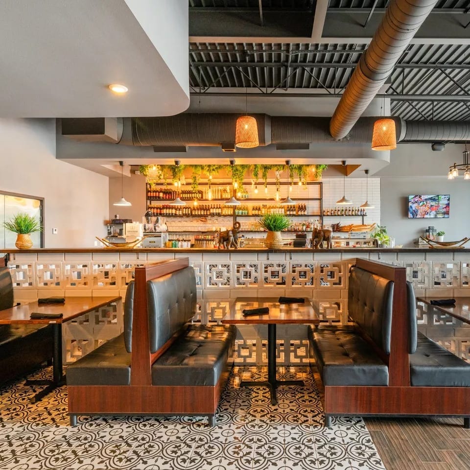 Restaurant interior with black leather booths, patterned floor, and a bar with hanging plants and bottles.