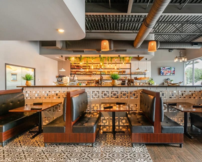 Restaurant interior with black leather booths, patterned floor, and a bar with hanging plants and bottles.