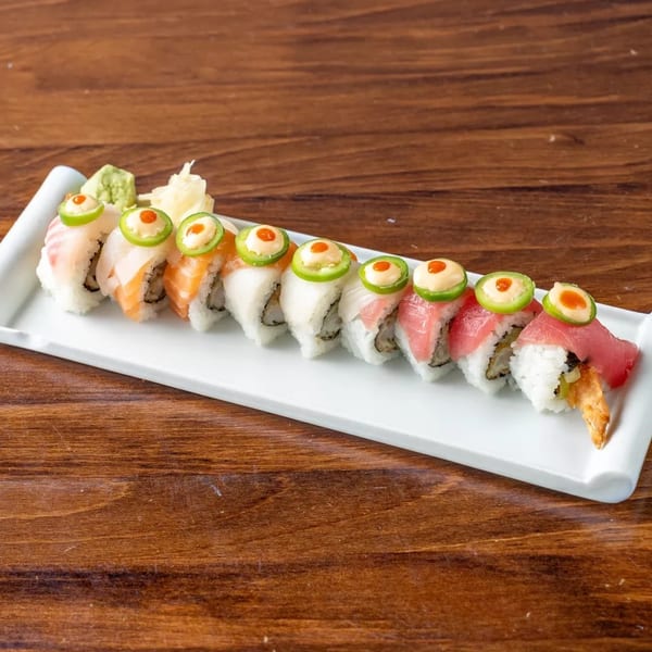 A long white plate of sushi rolls topped with fish, jalapeños, and sauces on a wooden table.