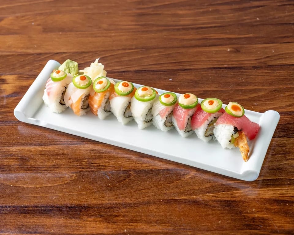 A long white plate of sushi rolls topped with fish, jalapeños, and sauces on a wooden table.