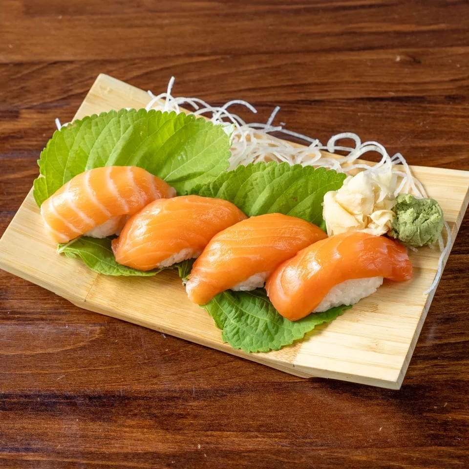 Four pieces of salmon nigiri sushi on green leaves, with ginger and wasabi, on a wooden board.
