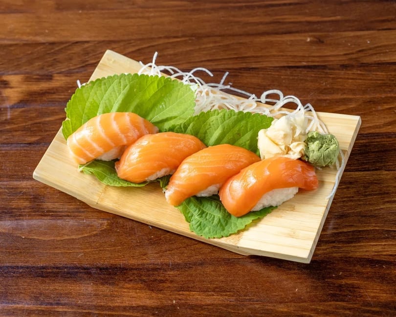 Four pieces of salmon nigiri sushi on green leaves, with ginger and wasabi, on a wooden board.