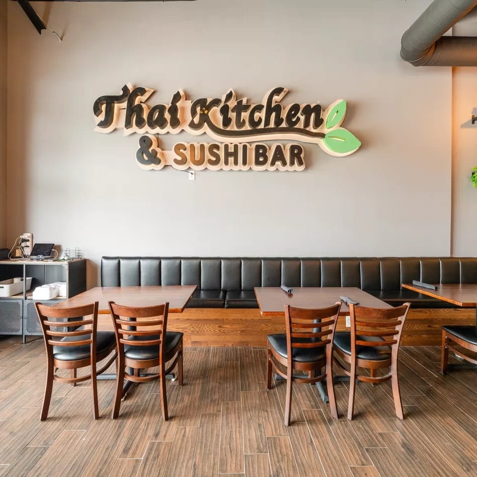 Interior of a Thai Kitchen & Sushi Bar restaurant with wooden tables, chairs, and a long black booth.