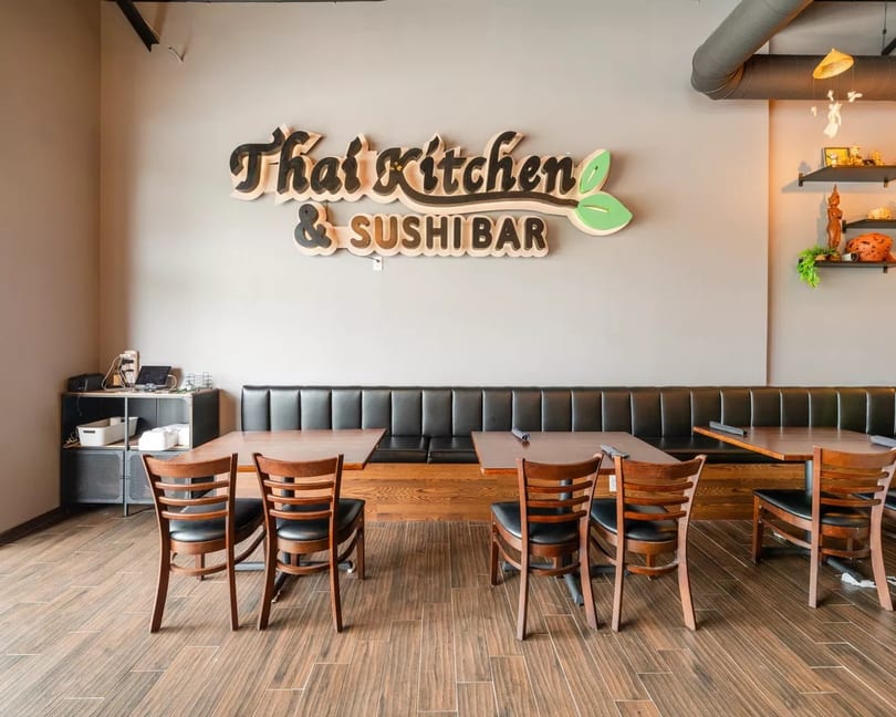 Interior of a Thai Kitchen & Sushi Bar restaurant with wooden tables, chairs, and a long black booth.